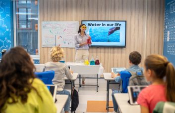 Chemistry lesson. Smiling woman in protective goggles holding flask with colored liquid standing looking at children in class