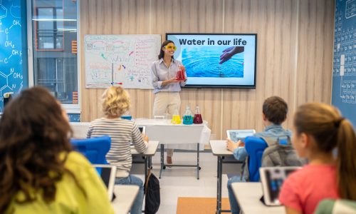 Chemistry lesson. Smiling woman in protective goggles holding flask with colored liquid standing looking at children in class